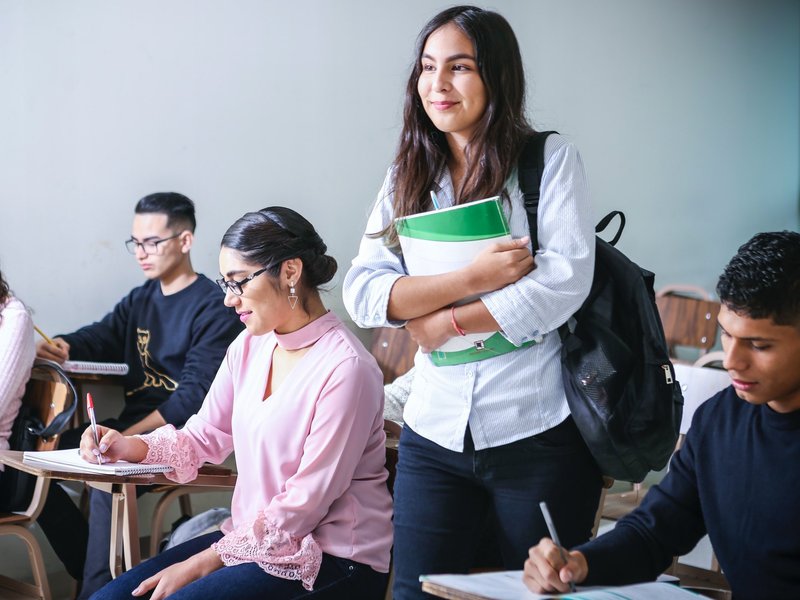 Eine Studentin hält Studienunterlagen im Arm und geht in einem Seminarraum, in dem bereits vier andere Student:innen sitzen, zu ihrem Platz.